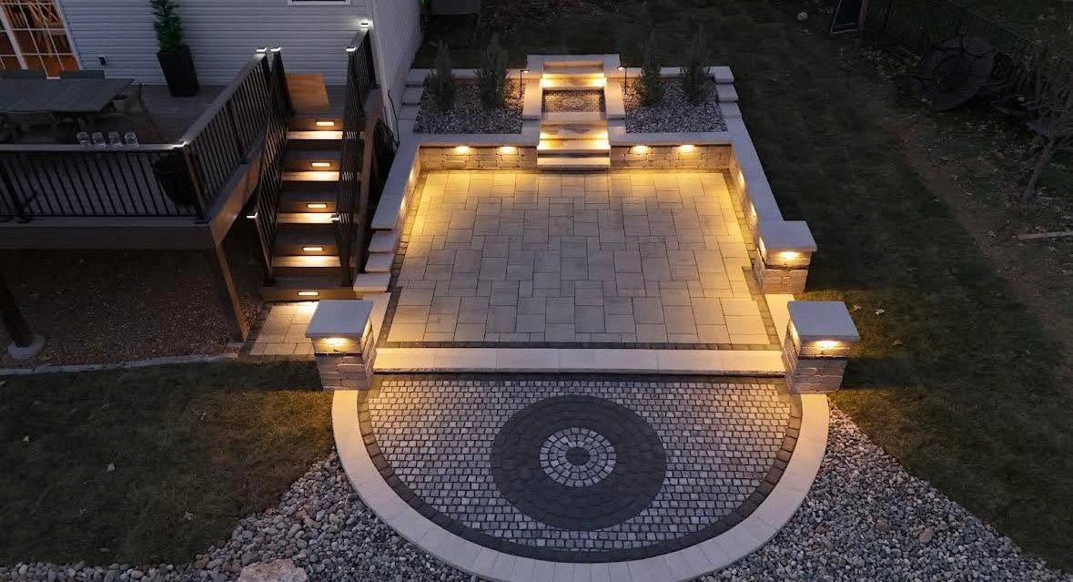 Lit paver patio with seat walls, steps, and a circular cobblestone inlay at night